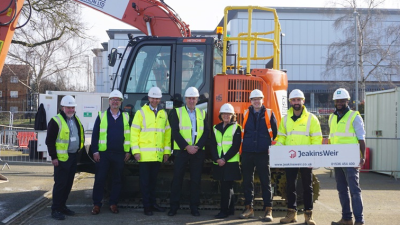 A group of people in high visibility jackets and white hard hats standing in front of a orange digger.