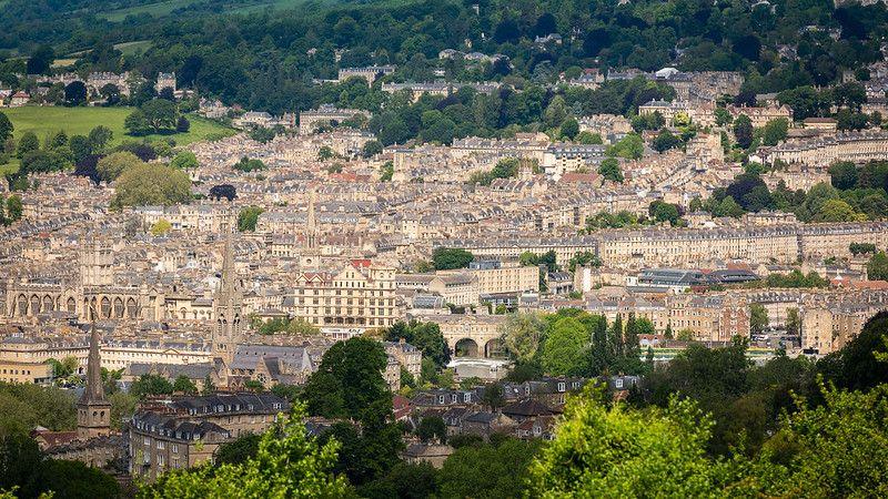 The city of Bath as captured from high up on a hill. You can see lots of ornate historic buildings interspersed with trees and green fields. 