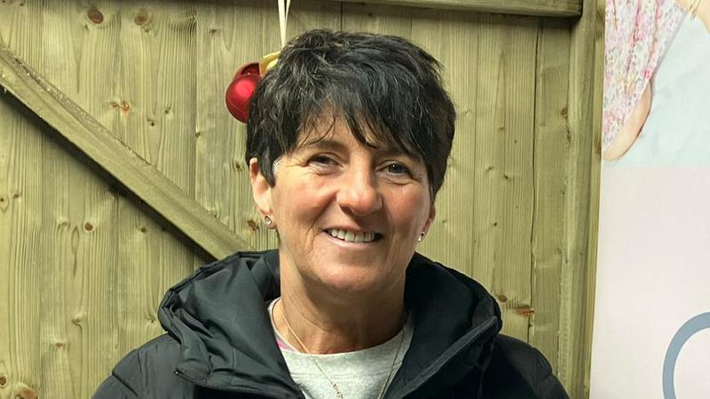 Denise McGilligan stands in front of a wooden door, with a sign promoting her wine business to her left. She has short dark hair and is wearing a heavy black winter coat, zipped up over a grey jumper.