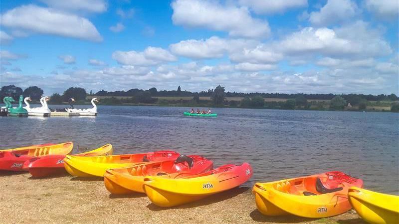 Seven canoes, which are yellow and red, and standing on a stretch of sand which goes into a lake. There are people in a green canoe on the water, and on the other side of the lake is a field. 