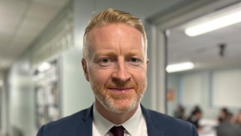 Brendan has short ginger hair, he has a gingery grey beard and is wearing a white shirt and maroon tie. He is wearing a navy blazer.