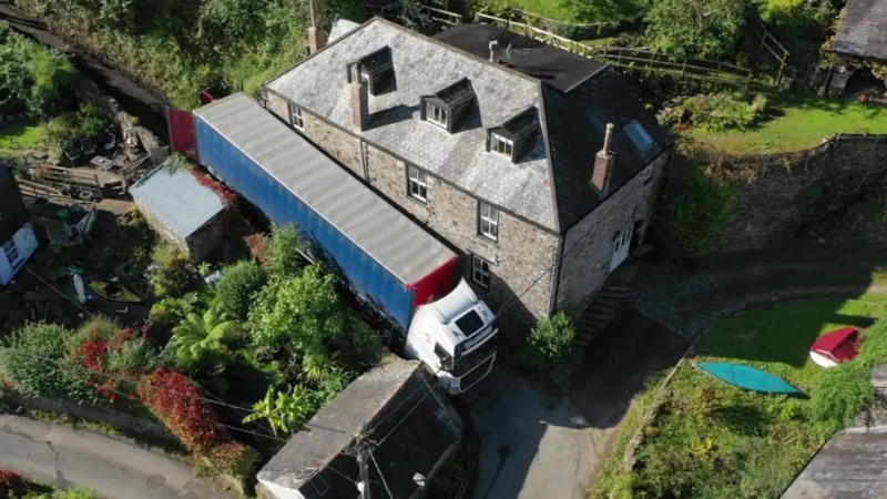 An aerial view of a blue, red and white lorry right up against a  
two-storey house made of stone. The surrounding area includes greenery, some scattered objects, and another small building with a black covering.