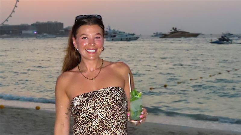 Isobel në plazh me një koktej, duke qëndruar përballë detit dhe një rrote të madhe. Është muzg.