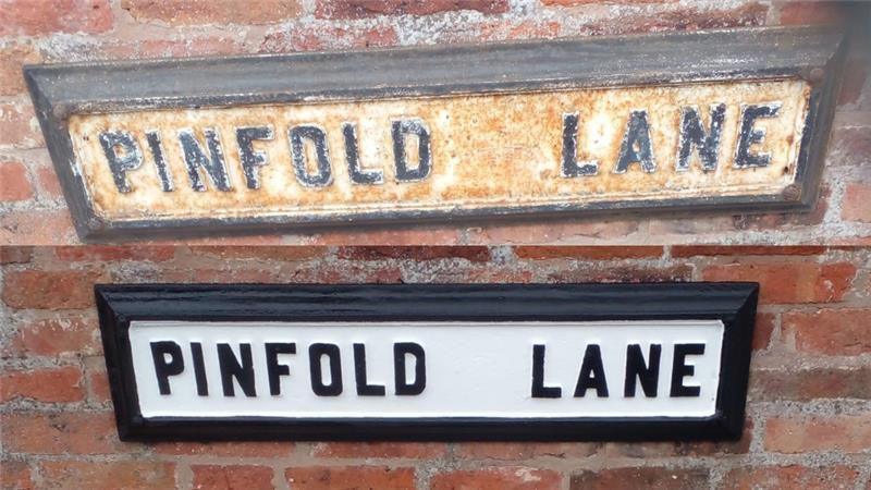 A rusting street sign that says 'Pinfold Lane' below it is the restored version with a white painted background and black lettering.