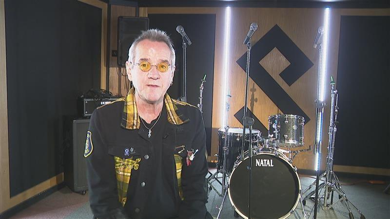 Stuart 'Woody' Wood stands in front of a drum kit. He has grey short hair with yellow tinted small round glasses. He wears a black jacket that has yellow tartan accents for the collar and pockets. 