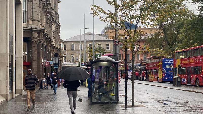 Belfast city centre, man walking away from the camera in grey trousers and black jacket holding a black umbrella. Another man walking towards the camera in tan coloured trousers and a grey and black stripes hoodie  