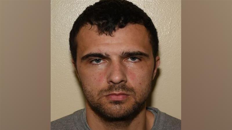 A police mugshot of Elias Morgan, who has black hair and dark stubble, staring into the camera with a blank expression.