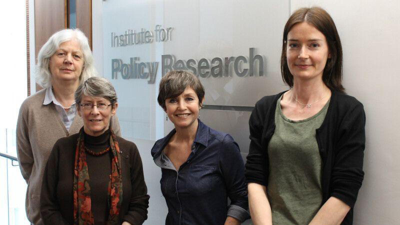 The UoB team looking into Universal Credit, from left to right: Professor Jane Millar, Fran Bennett, Rita Griffiths and Marsha Wood. They are all wearing shirts with blouses, and one is wearing an orange and brown scarf. They are leaning on a wall where a big glass sign reads 'Institute for Policy Research'.