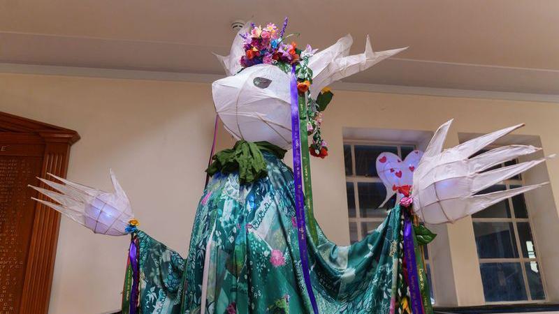 A large willow puppet covered in tissue paper like lantern. It has a head a bit like a deer and large lantern hands, wearing a colourful floral headdress and a green robe.