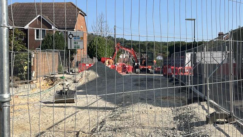 A work site with a red digger at the far end. The site is filled with dirt and gravel. The site is blocked off by a metal railed fence.