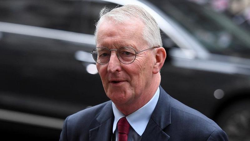 Hilary Benn - a white haired man with black oval glasses looking wearing a navy suit, light blue shirt and red tie. 