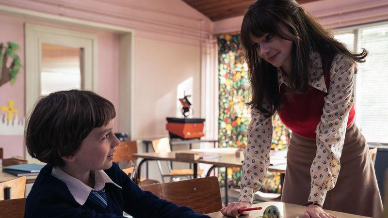 a child in a school uniform with a big shirt collar, sitting at a table and looking up at a smiling woman who is leaning on the table
