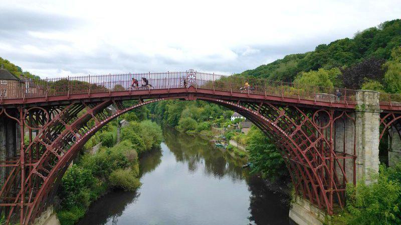 A wide angle of the red, metal Ironbridge, which stands above the River Severn. There are three cyclists riding over the bridge. On either side of the river are green trees and bushes, as well as a small white house on the banks of the river.