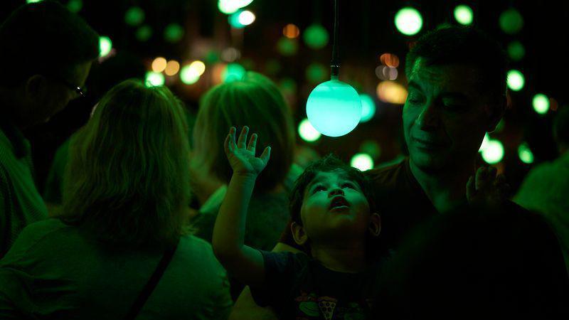 A small child, held by an adult, looks up in amazement at a blue glowing bulb as part of an art installation. There are other people out of focus in the background, also looking up at dozens of blue, green and orange glowing bulbs suspended from the ceiling.