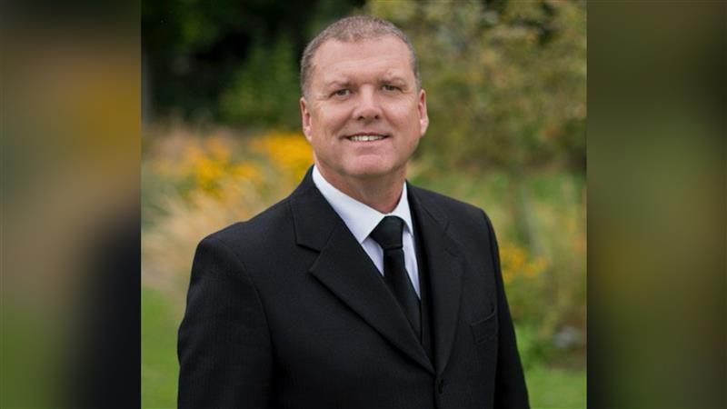 A man in a black funeral suit and tie. The image is taken outdoors in a park or garden, but the background is out of focus.