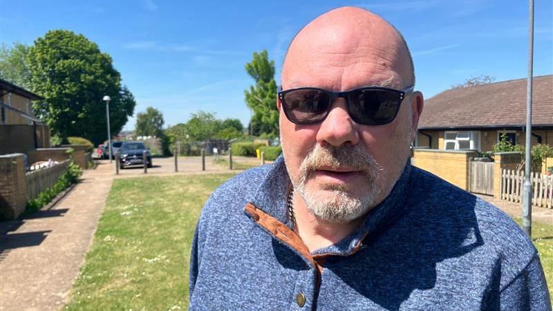 A bald man with dark glasses and a goatee wearing a blue jumper looks at the camera.