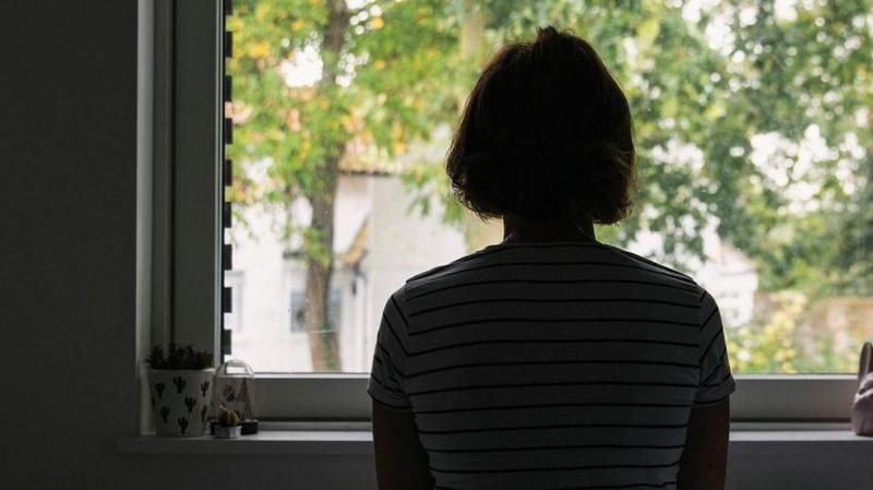 A woman sat in front of a window with her back to the camera. She has bobbed hair and is wearing a striped T-shirt.
