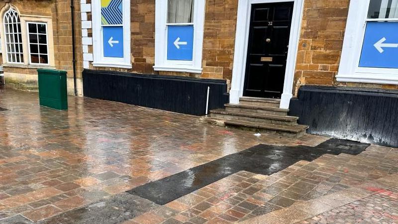 A long, black asphalt strip strip runs over some new-looking brick paving. The paving is in front of an old, stone building with a black door. 