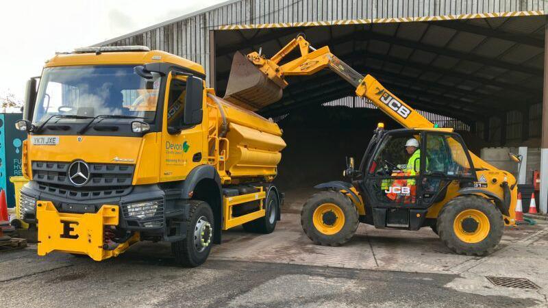 A yellow lorry is parked outside a gritting barn as a JCB digger loads it with grit.