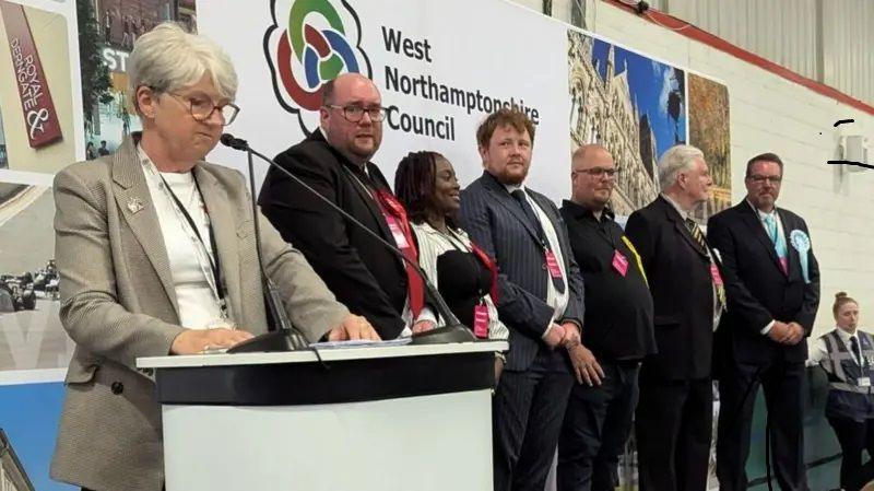 Anna Earnshaw with short white hair and glasses wearing a light top and beige jacket. She is standing behind a lectern with a microphone. Election candidates are standing to her left - five men in dark jackets and ties and one woman in a light-coloured jacket. They are standing in front of a West Northamptonshire Council banner at an election count.