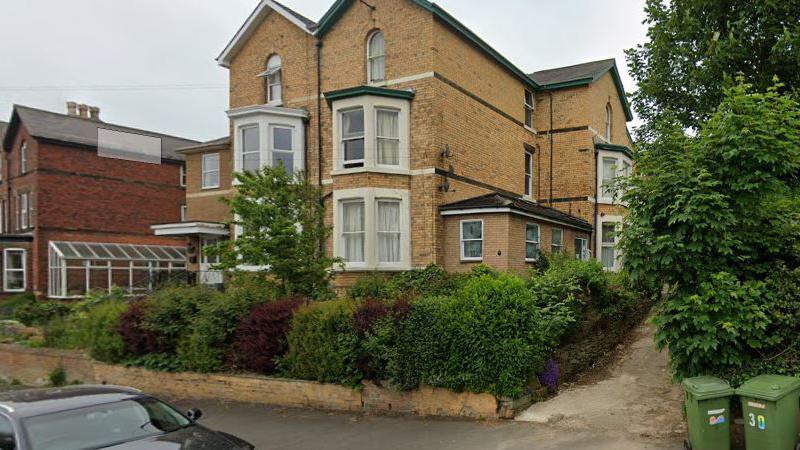 A large brick house surrounded by privet hedge and shrubs with large bay windows.