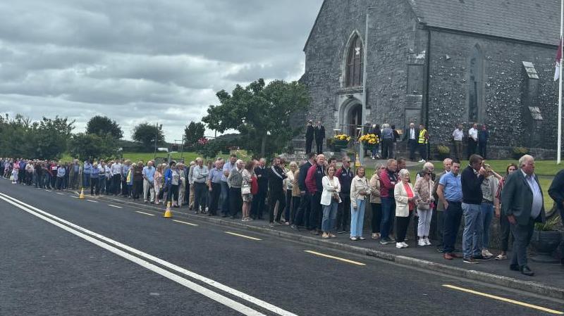 Many people lined up on a street outside a church. There is a cloudy sky above. 