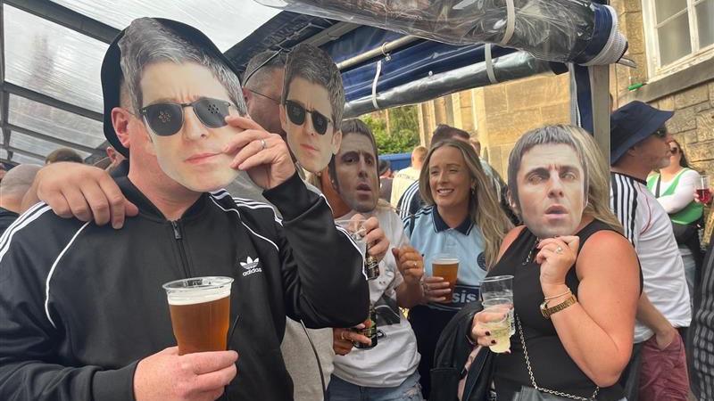 A group of Oasis fans. A man on the near left is wearing a black adidas track top with a white logo on the upper right chest and three white stripes on the arms. He is holding a face mask of Noel Gallagher to his face in his left hand. A man next to him has the same mask attached to his face. On the far side, a woman holding a cup of beer is wearing an oasis T-shirt. A woman next to her wearing a black top is holding a cup of wine in one hand and a face mask of Liam Gallagher to her face with the other.