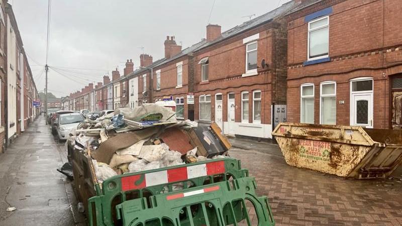 Street lined with small semi detached houses with skips on the side of the road