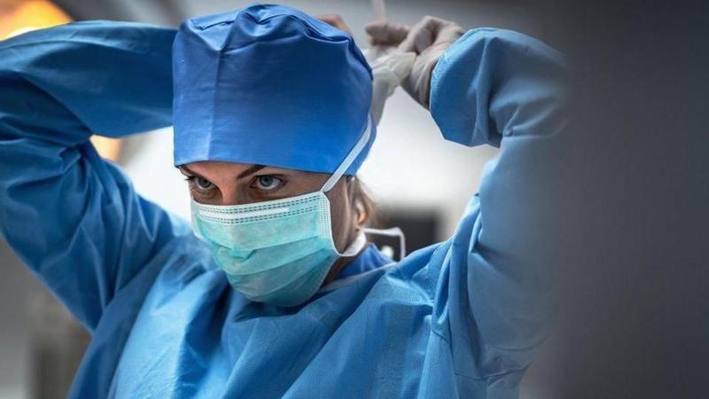 A surgeon in blue scrubs putting on a white face mask