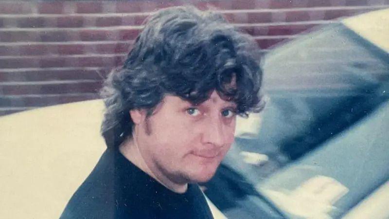 A man with long, wavy black hair looking at the camera. He is wearing a black tshirt. He is stood by the front of a white car