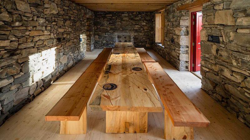 A long wooden table inside the cottage. There is stone brick work along all four walls, with the red door open allowing in the daylight. Two long wooden benches are on either side of the table. 