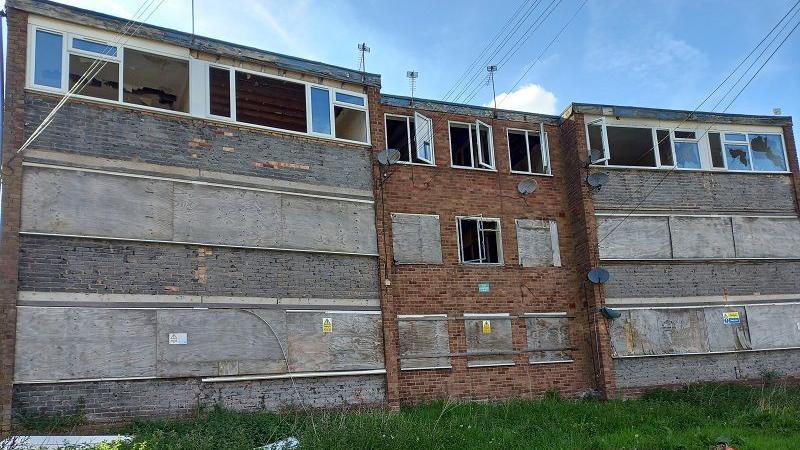 A run-down block of flats. It is a grey and red brick building that looks old. There are windows on the second floor that are open, but windows on the ground and first floor are boarded up with wood. Some of the windows are smashed.