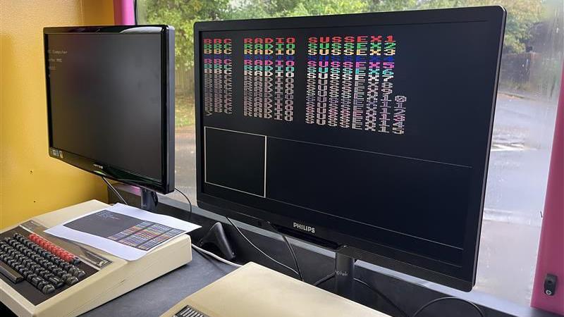 A computer screen which reads BBC Radio Sussex in multi colours. It was coded on a BBC Microcomputer which is a computer system with a keyboard from 1991 which has black and red buttons and a cream casing