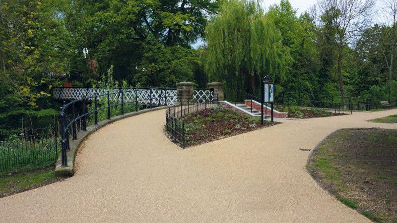 An ornate bridge in Pump Room gardens. Honey-coloured gravel paths lead to the bridge, with black iron railings on the approach. Planted beds and grass are also visible 