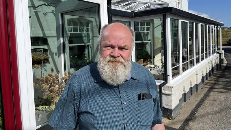 Philip Harris, a middle aged man with a bushy grey beard, wears a blue sleeveless shirt and stands outside the white-fronted Sportsman pub in Seasalter.