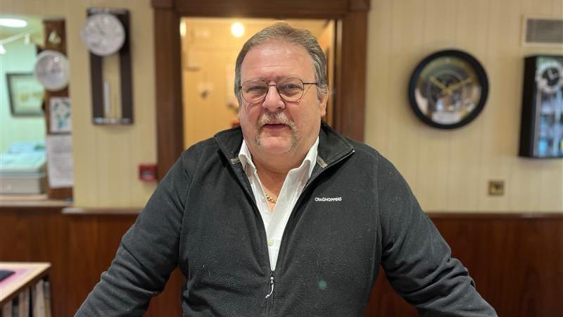 Will Van Zanten, Owner of Machon's Jewellers. He has grey hair and a moustache. He is wearing round glasses, a white shirt and a black zip-up fleece, a gold chain is peeking through. His arms a leaning on his desk outstretched and he is looking directly at the camera. 