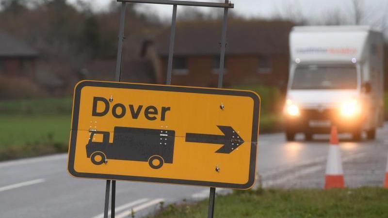 A sign for Dover: a rectangular yellow road sign sitting on a grey stand. The word "Dover" is written on the sign in black. A black arrow pointing right and a black image of a lorry can also be seen on the sign. The sign is sitting on the right-hand side of a black tarmac road. A white van with its lights on can be seen in the background.