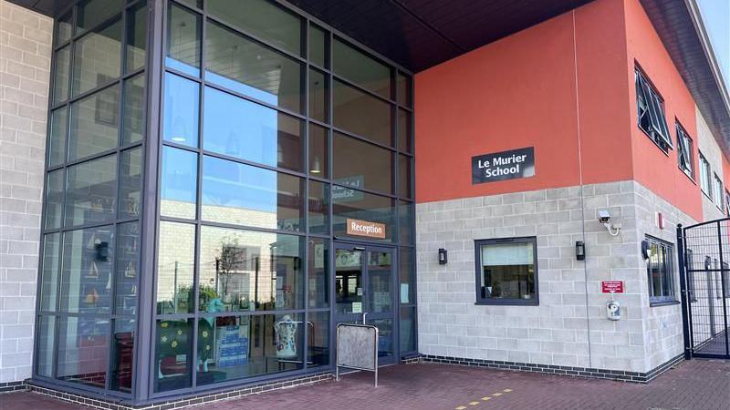 A picture of the outside of the school which shows the reception with big glass windows. The remainder of the building is brick with the top half painted red.