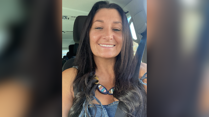 Paula has taken a selfie in her car. She is smiling at the camera and has chest length black hair.
