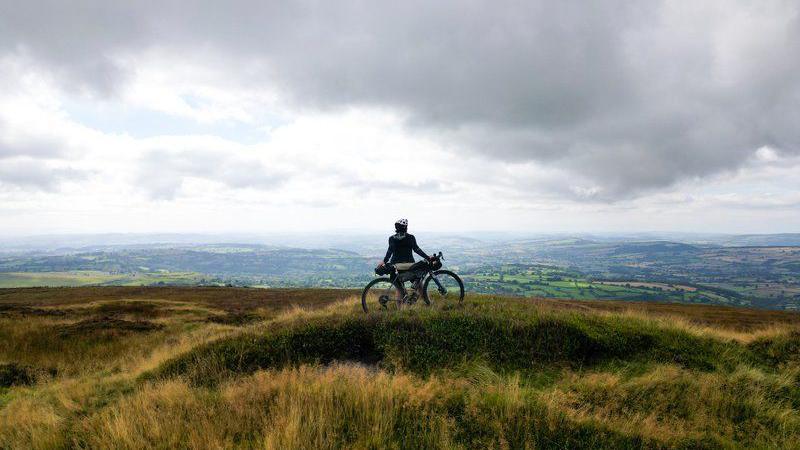 A cyclist is standing in a field, leaning against their bike. They have a helmet on. They are looking out on a vast view of the horizon, which includes miles of green fields and hills in the distance. The sky is bright and grey, with white clouds that create a slight mist in the distance.