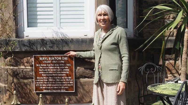 A person stands outside a house beside a memorial plaque mounted on a stone wall. The plaque commemorates cyclist “Beryl Burton OBE,” with engraved details about achievements and years (1937–1996). Garden furniture and plants are visible nearby, with bright daylight illuminating the scene.