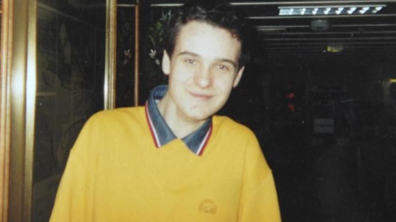 Alan Radford with black, curly hair, smiles at the camera wearing a yellow sweat shirt with a grey, red and white collar.