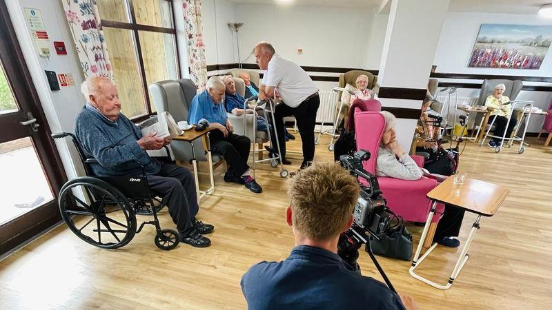 The picture features a care home setting where a man in a blue shirt is with a camera and recording care home residents. In the picture, an elderly man in a wheelchair wearing a blue jumper. Beside him is a row of elderly residents and a staff member talking. 
To the right of the camera man, a woman is sitting in a pink chair, with her head leaning on her fist