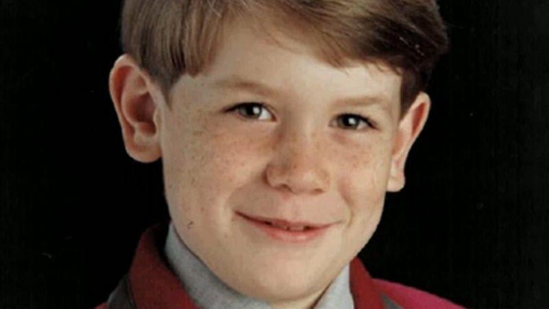 James Barker has brown hair parted to the side and brown eyes. He is smiling to the camera while wearing a burgundy coloured school blazer