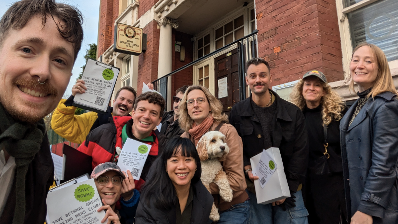 A group of men and women smiling at the camera holding up A4 pieces of paper that say 'Save BGWMC'. One person is holding a fluffy white dog in the middle of the group. 