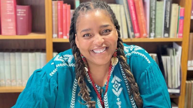 Désirée Reynolds has long, brown braided hair and is wearing a turquoise top with white stitching across her shoulders and down her chest. She is smiling at the camera as she sits in front of a bookshelf.