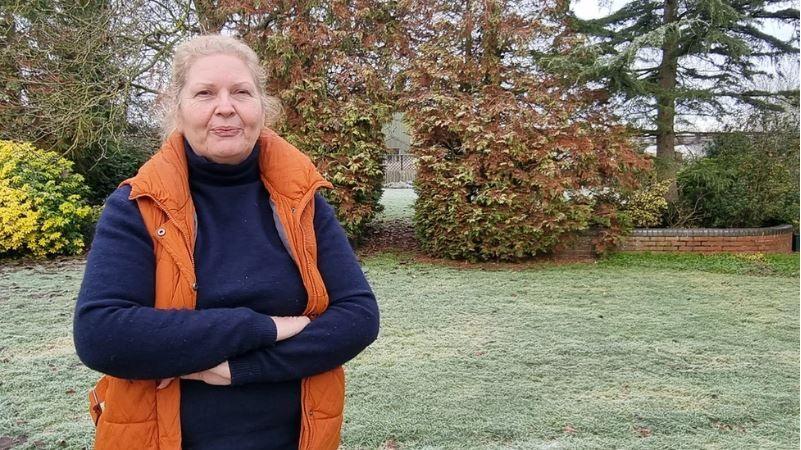 Sue Butler with tied back blonde hair, wearing a navy polo neck and orange body warmer, standing with her arms crossed in a garden covered in frost.