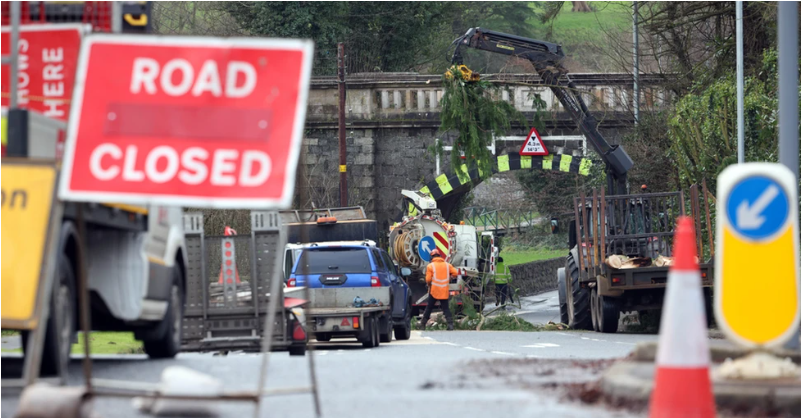 A shot of works being carried out on a road where branches of a tree have fell on the road. There is a red 'road closed' sign and workers with a blue pick-up truck, a tractor and trailer.