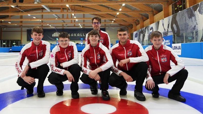 A team picture of England's men's junior curling team on the ice rink, with five crouching in front and another player in the middle behind. They are all wearing black trousers and shoes, and red tracksuit tops with white arms.