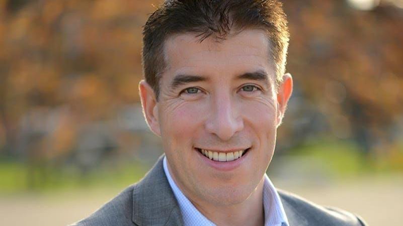 A close-up image of Michael Moran, wearing a grey suit and blue shirt. He has short brown hair and is smiling.